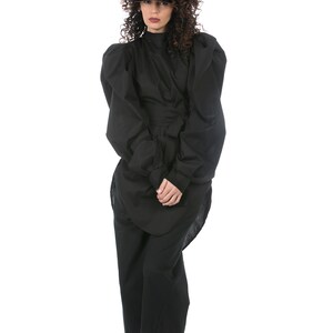May include: A full-length shot of a person wearing a black outfit. The outfit includes a high-neck top with large, puffed sleeves, a wrap-around waist detail, and wide-leg pants. White sneakers complete the look. The person has curly brown hair.