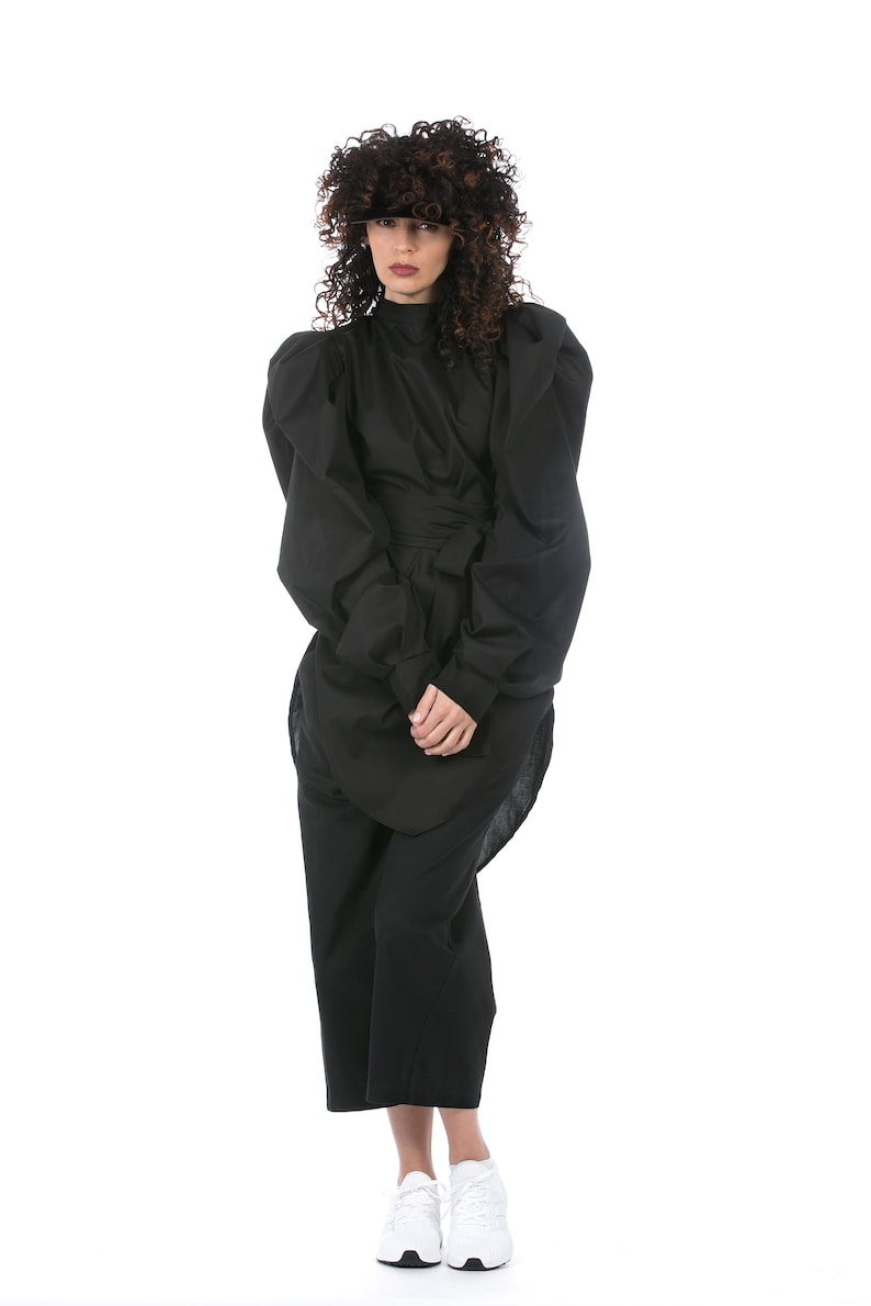 May include: A full-length shot of a person wearing a black outfit. The outfit includes a high-neck top with large, puffed sleeves, a wrap-around waist detail, and wide-leg pants. White sneakers complete the look. The person has curly brown hair.