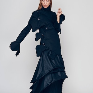 May include: A woman wearing a black, asymmetrical, long-sleeved dress with a high neck and multiple layers. The dress has a unique design with several fabric panels and buckles.