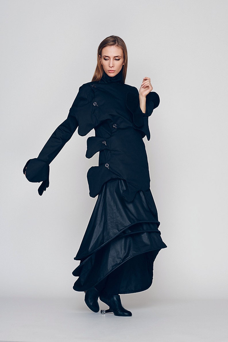 May include: A woman wearing a black, asymmetrical, long-sleeved dress with a high neck and multiple layers. The dress has a unique design with several fabric panels and buckles.