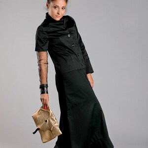 May include: A black outfit consisting of a short-sleeved button-up top and a long skirt. The outfit is paired with a gold clutch. The model is also wearing a black cuff bracelet and a sheer sleeve.