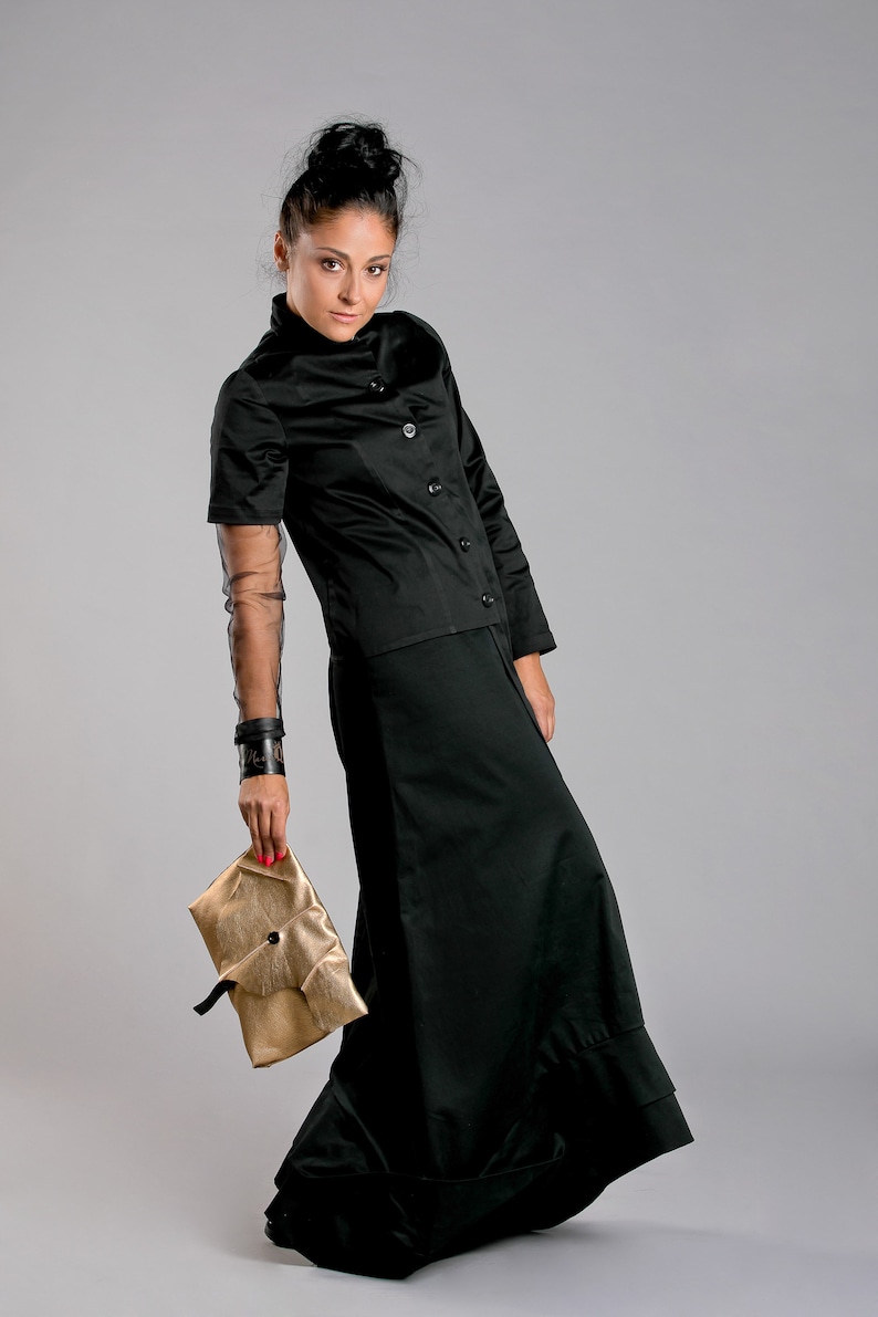 May include: A black outfit consisting of a short-sleeved button-up top and a long skirt. The outfit is paired with a gold clutch. The model is also wearing a black cuff bracelet and a sheer sleeve.