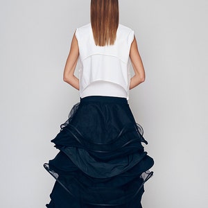 May include: A woman wearing a white sleeveless top and a black tulle skirt with multiple layers. The skirt is gathered and ruffled, creating a voluminous and dramatic look.