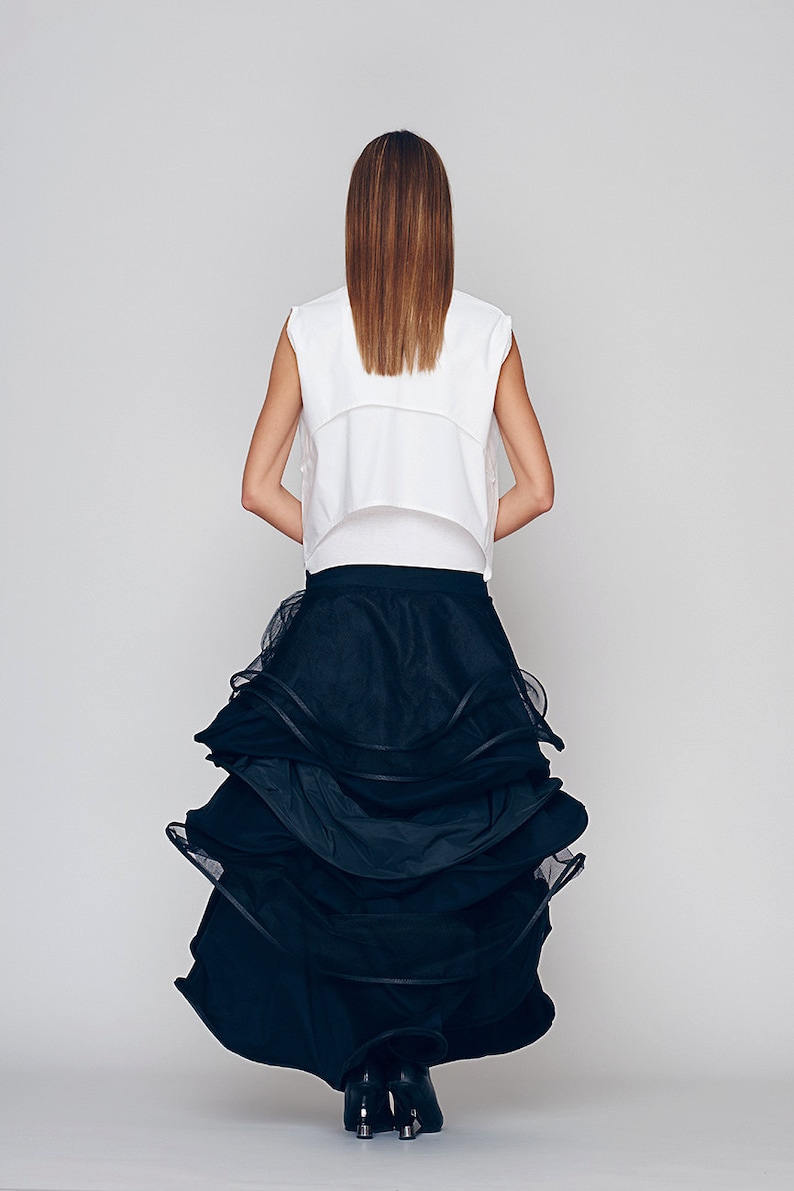May include: A woman wearing a white sleeveless top and a black tulle skirt with multiple layers. The skirt is gathered and ruffled, creating a voluminous and dramatic look.