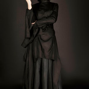 May include: A woman wearing a black, long-sleeved dress with a high neck and a flowing skirt. The dress has a unique design with layers and a belt.