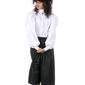 May include: A woman wearing a white button-up shirt with puffed sleeves and a black skirt. She is wearing white sneakers.