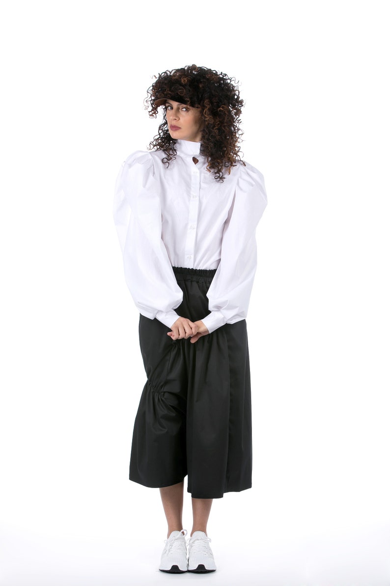 May include: A woman wearing a white button-up shirt with puffed sleeves and a black skirt. She is wearing white sneakers.