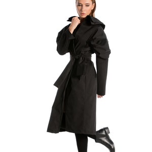 May include: A woman wearing a black long-sleeved coat with a belt and a hood. She is wearing black boots and has her hair down.