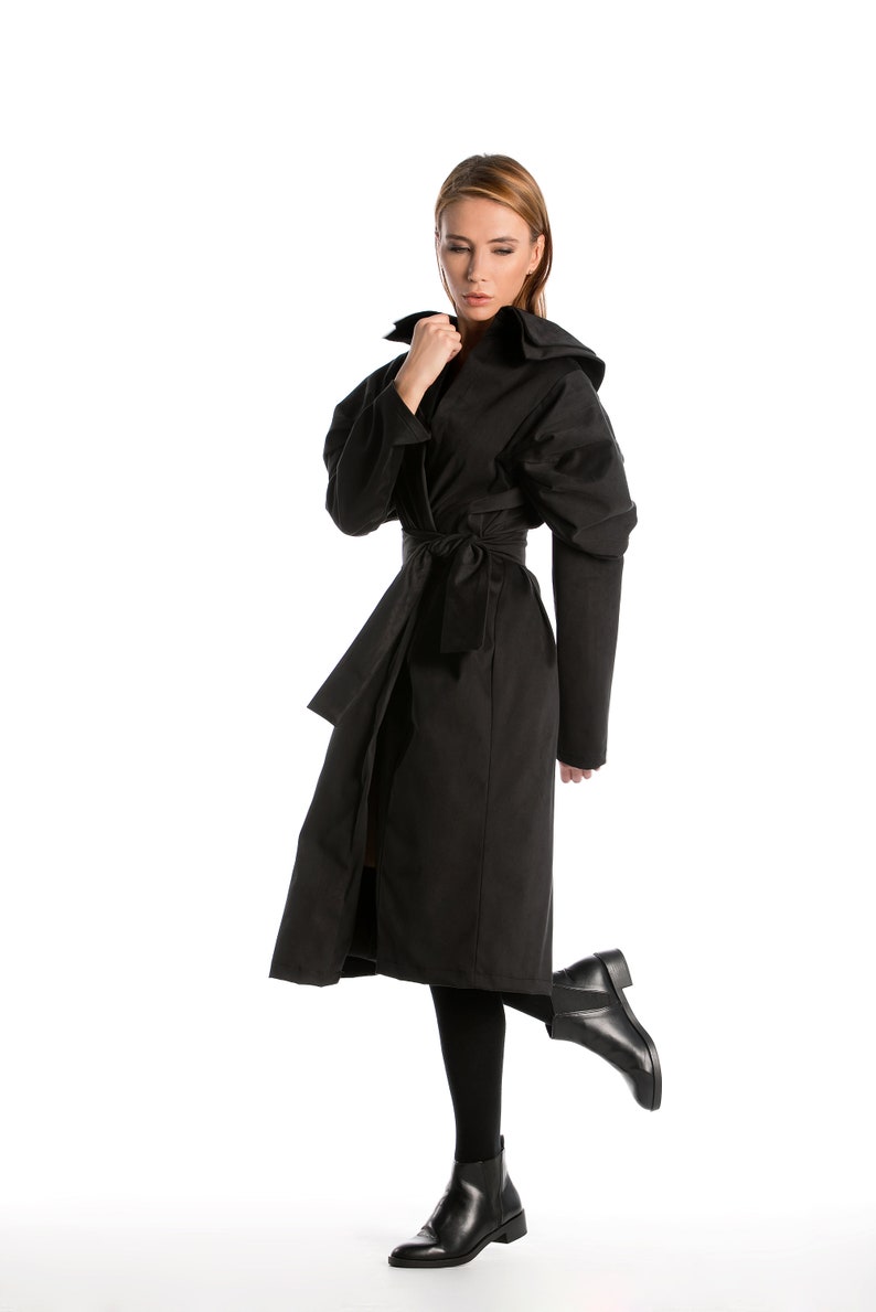 May include: A woman wearing a black long-sleeved coat with a belt and a hood. She is wearing black boots and has her hair down.