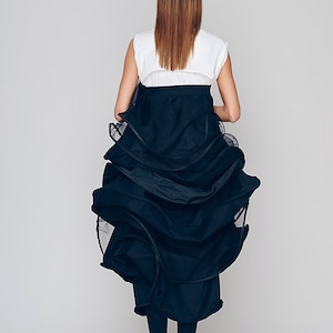 May include: A woman wearing a white sleeveless top with a black belt and a black layered skirt. The skirt is made of a sheer fabric and has a ruffled design.