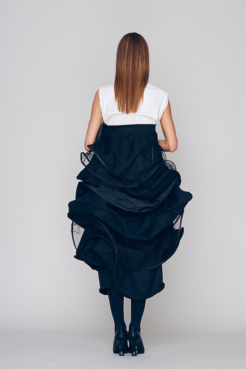 May include: A woman wearing a white sleeveless top with a black belt and a black layered skirt. The skirt is made of a sheer fabric and has a ruffled design.