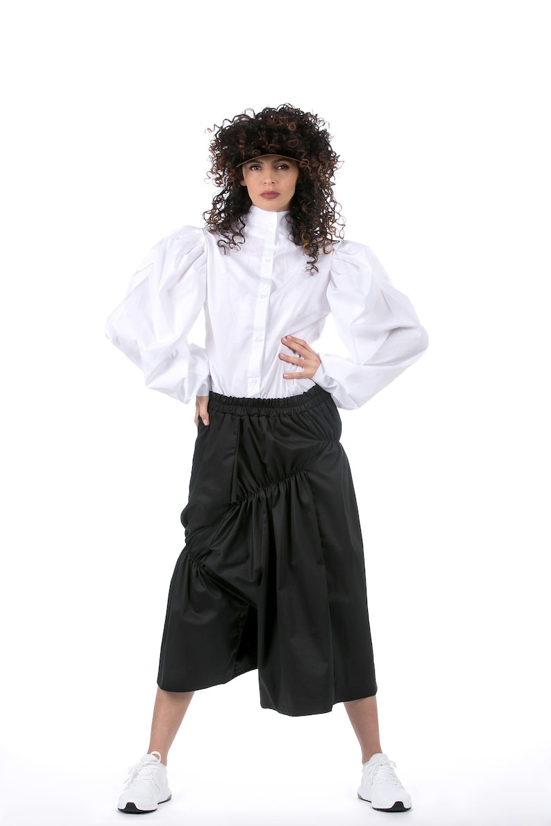 May include: A woman wearing a white button-up shirt with puffed sleeves and a black gathered skirt. She is wearing white sneakers and has curly brown hair.