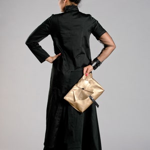 May include: A woman in a black outfit, including a long skirt and jacket, holds a gold clutch. The jacket has sheer sleeves. The outfit is set against a gray background.