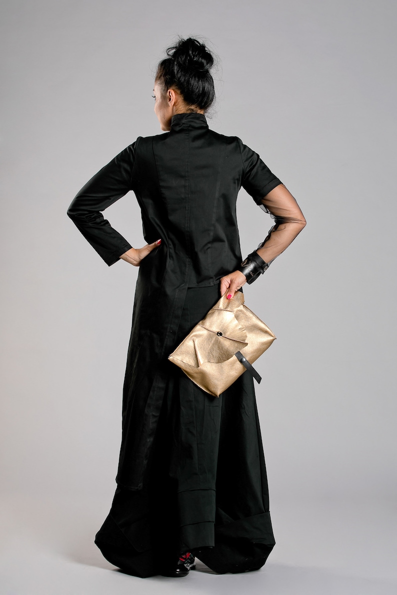 May include: A woman in a black outfit, including a long skirt and jacket, holds a gold clutch. The jacket has sheer sleeves. The outfit is set against a gray background.