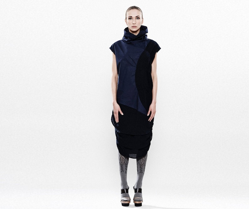 May include: A woman wearing a black and blue sleeveless dress with a high neck and a geometric pattern. She is wearing gray patterned tights and black heeled shoes.