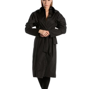 May include: A woman wearing a black long-sleeved coat with a hood and a belt. She is standing with her arms crossed and looking down.