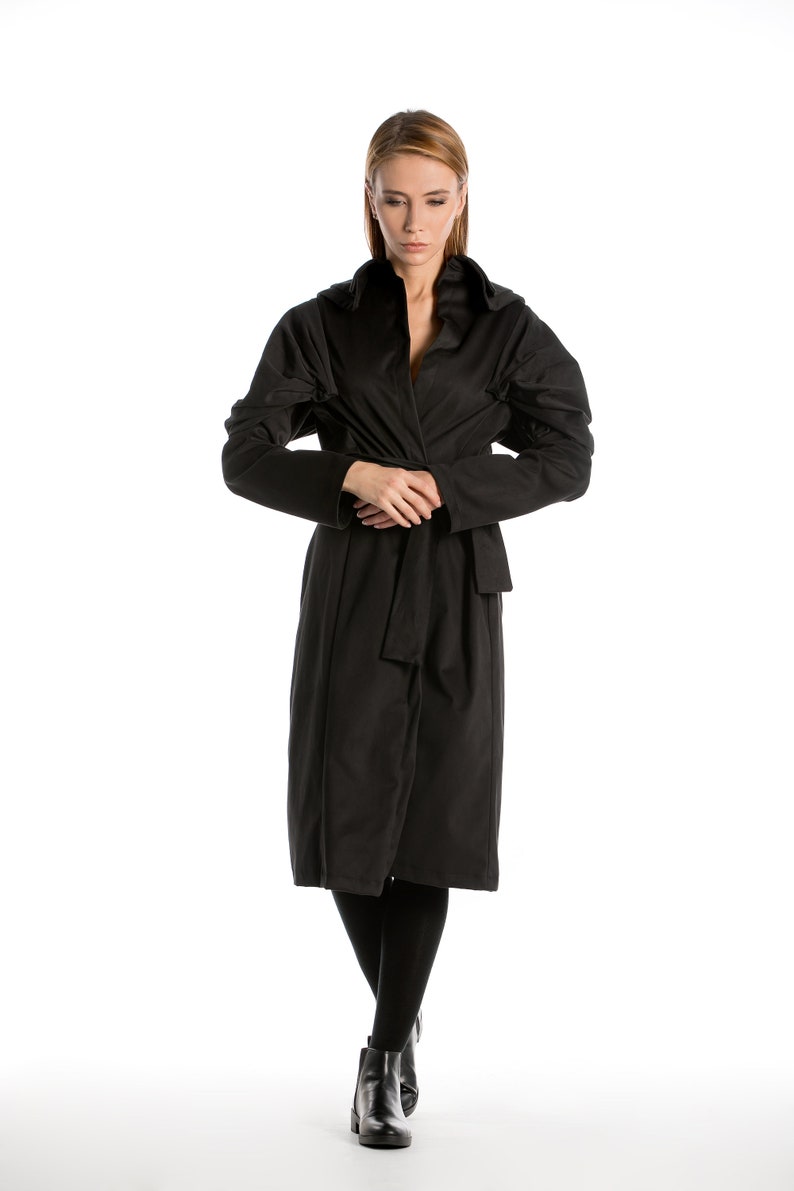 May include: A woman wearing a black long-sleeved coat with a hood and a belt. She is standing with her arms crossed and looking down.