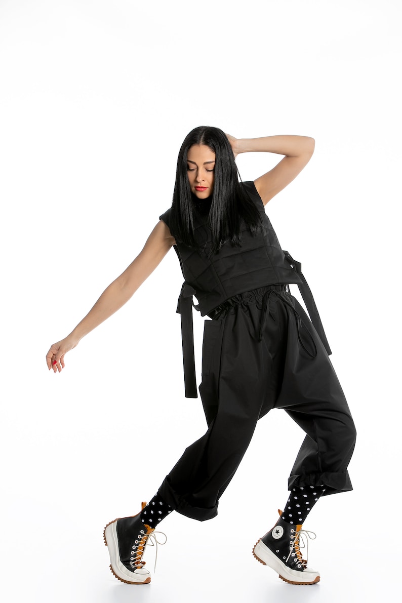 May include: A woman wearing a black sleeveless top with a quilted design and black trousers. She is wearing black and white polka dot socks and black and white high-top trainers with brown accents.