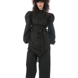 May include: A black outfit featuring a high-necked top with voluminous sleeves, a wrap-style skirt, and wide-leg pants. The outfit is cinched at the waist with a matching belt. White sneakers complete the look. The model is wearing a dark wig.