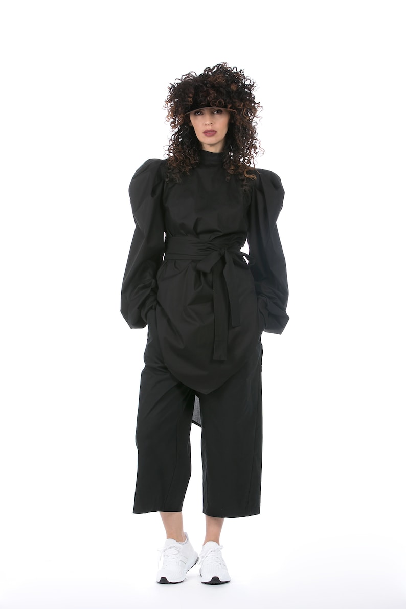 May include: A black outfit featuring a high-necked top with voluminous sleeves, a wrap-style skirt, and wide-leg pants. The outfit is cinched at the waist with a matching belt. White sneakers complete the look. The model is wearing a dark wig.