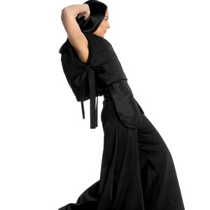 May include: A black outfit consisting of a quilted top with bow detail and wide-leg pants. The top has a unique design with a cape-like element. The model is wearing white sneakers. The outfit is a modern take on a classic style.