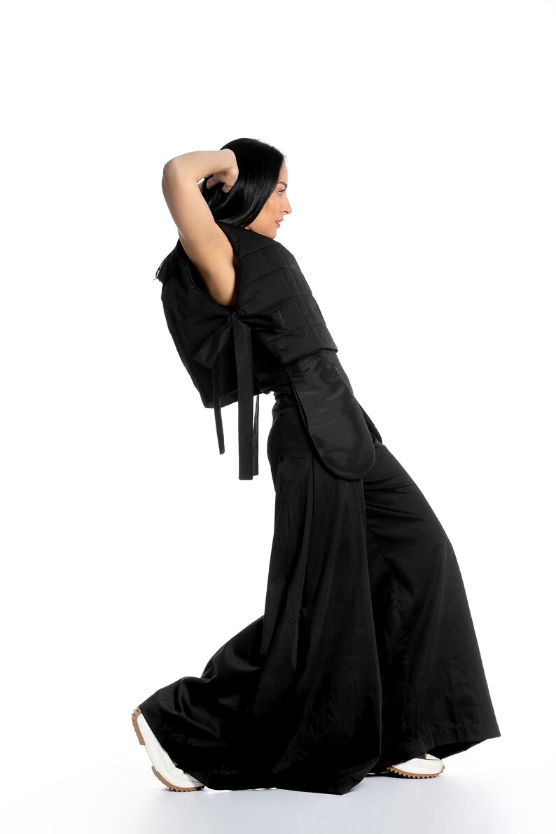 May include: A black outfit consisting of a quilted top with bow detail and wide-leg pants. The top has a unique design with a cape-like element. The model is wearing white sneakers. The outfit is a modern take on a classic style.