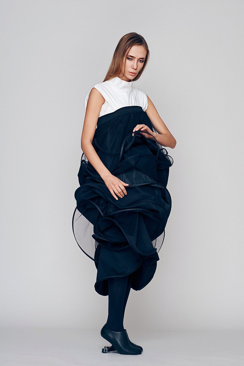 May include: A woman wearing a white sleeveless top and a black, voluminous skirt with a ruffled hem. She is wearing black tights and black shoes with a unique design.