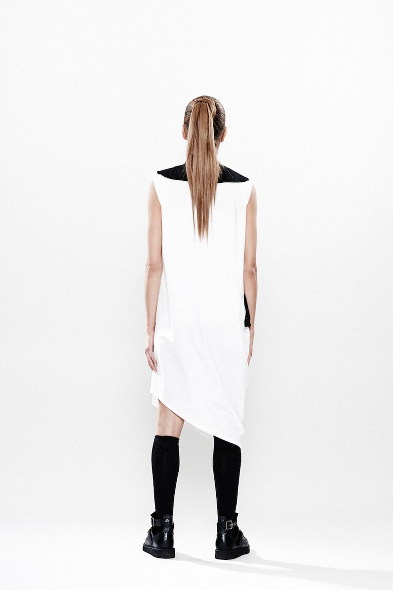 May include: A white sleeveless dress with a black collar and a black panel on the back. The dress has a loose fit and a slightly asymmetrical hemline. The model is wearing black knee-high socks and black shoes.