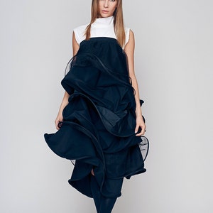 May include: A woman wearing a black dress with a white top and black tights. The dress is made of a layered fabric that creates a dramatic and flowing silhouette. She is wearing black ankle boots.