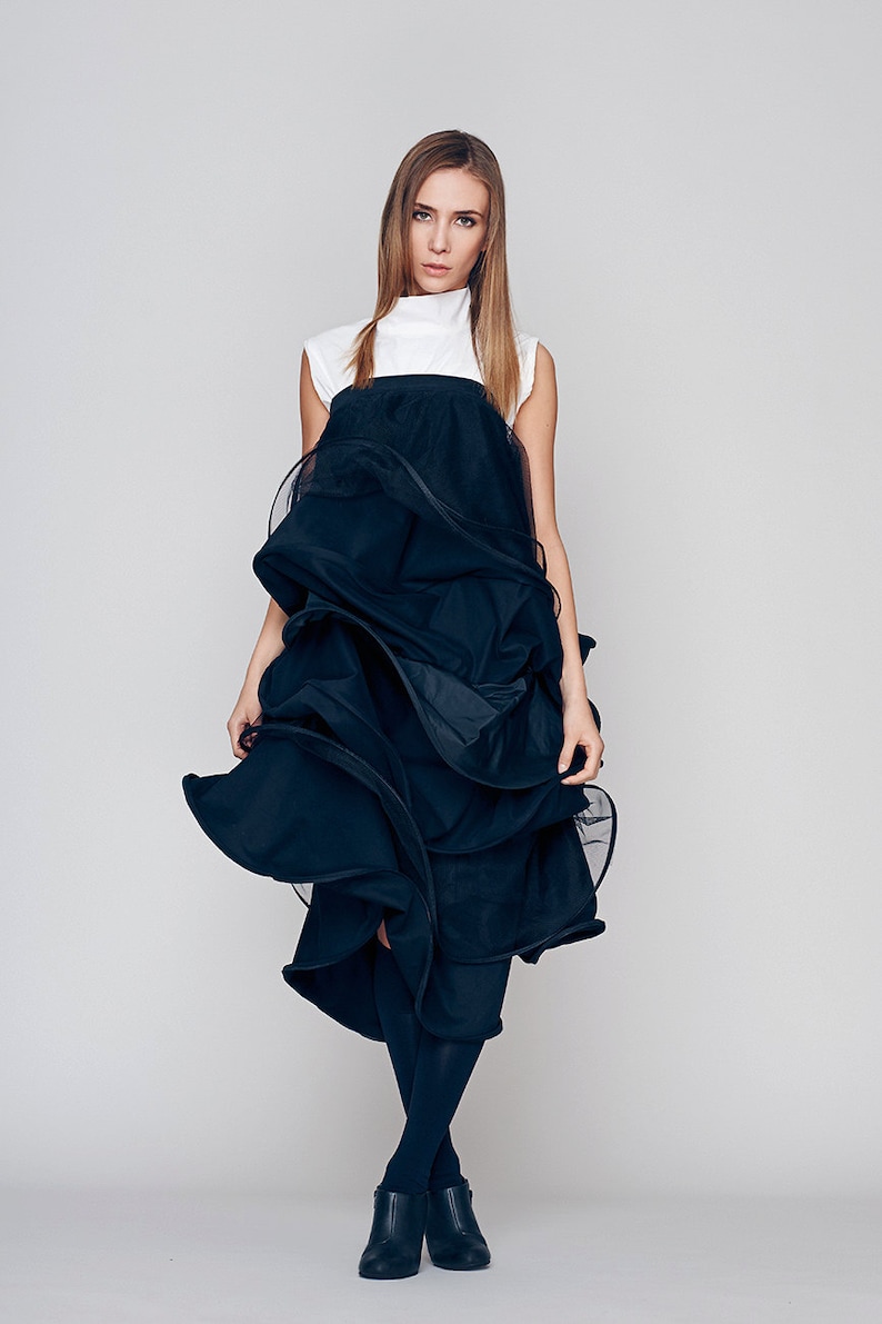 May include: A woman wearing a black dress with a white top and black tights. The dress is made of a layered fabric that creates a dramatic and flowing silhouette. She is wearing black ankle boots.
