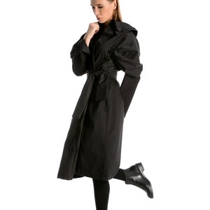 May include: A woman wearing a black long-sleeved coat with a hood and a belt. She is wearing black tights and black ankle boots. She is standing on one leg with her other leg bent at the knee.