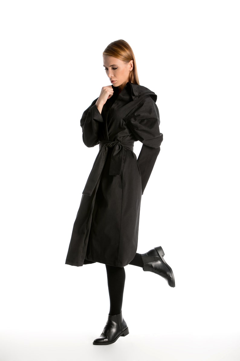 May include: A woman wearing a black long-sleeved coat with a hood and a belt. She is wearing black tights and black ankle boots. She is standing on one leg with her other leg bent at the knee.