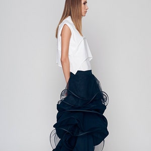 May include: A woman wearing a white sleeveless top and a black tulle skirt with multiple layers. The skirt is gathered and ruffled, creating a voluminous and dramatic look.