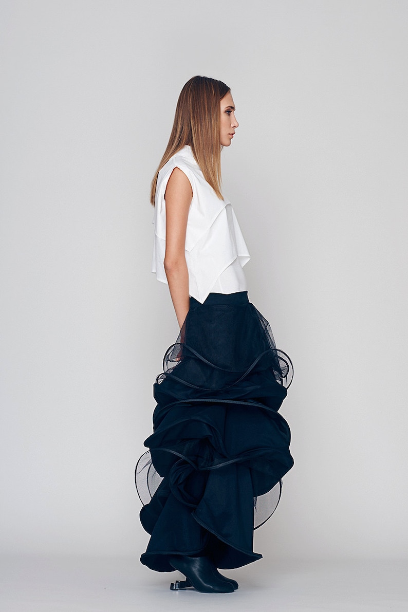 May include: A woman wearing a white sleeveless top and a black tulle skirt with multiple layers. The skirt is gathered and ruffled, creating a voluminous and dramatic look.