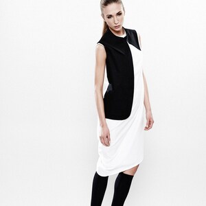 May include: A woman wearing a white dress with a black sleeveless vest. She is wearing black socks and black shoes.