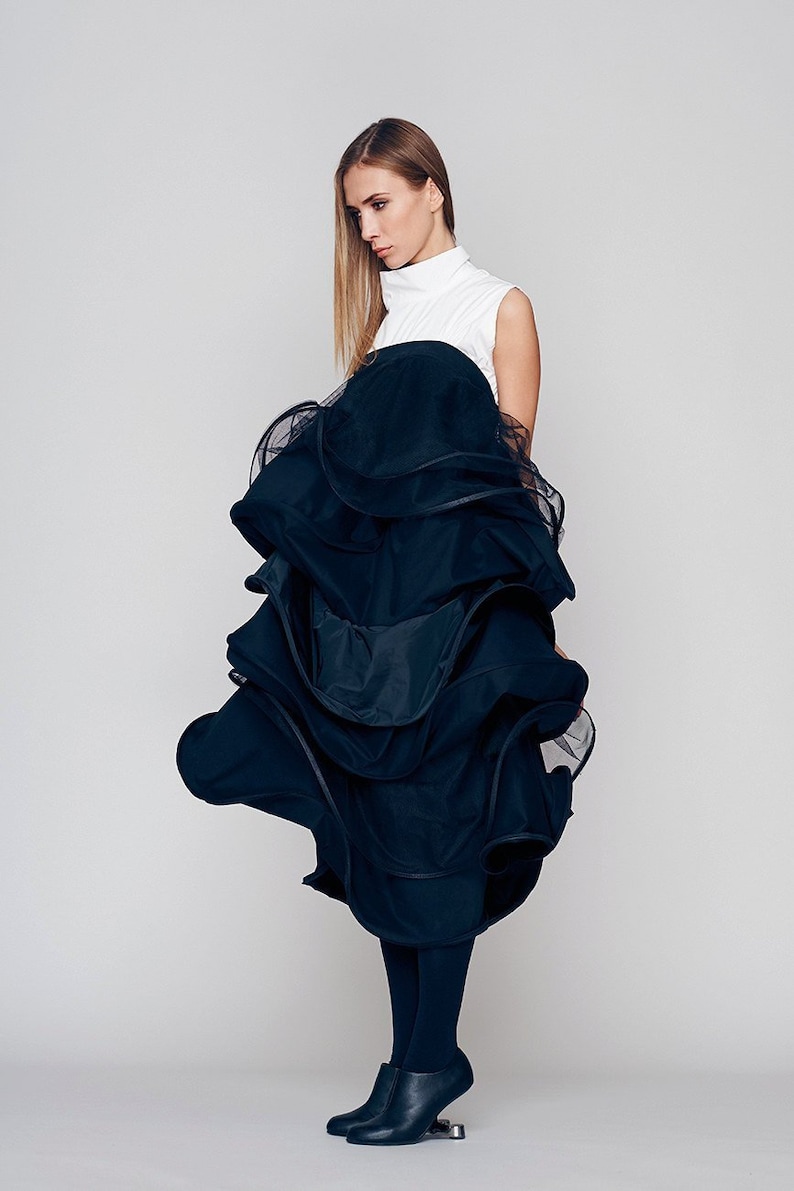 May include: A woman wearing a white sleeveless top and a black layered skirt with a ruffled hem. She is wearing black tights and black heeled shoes.