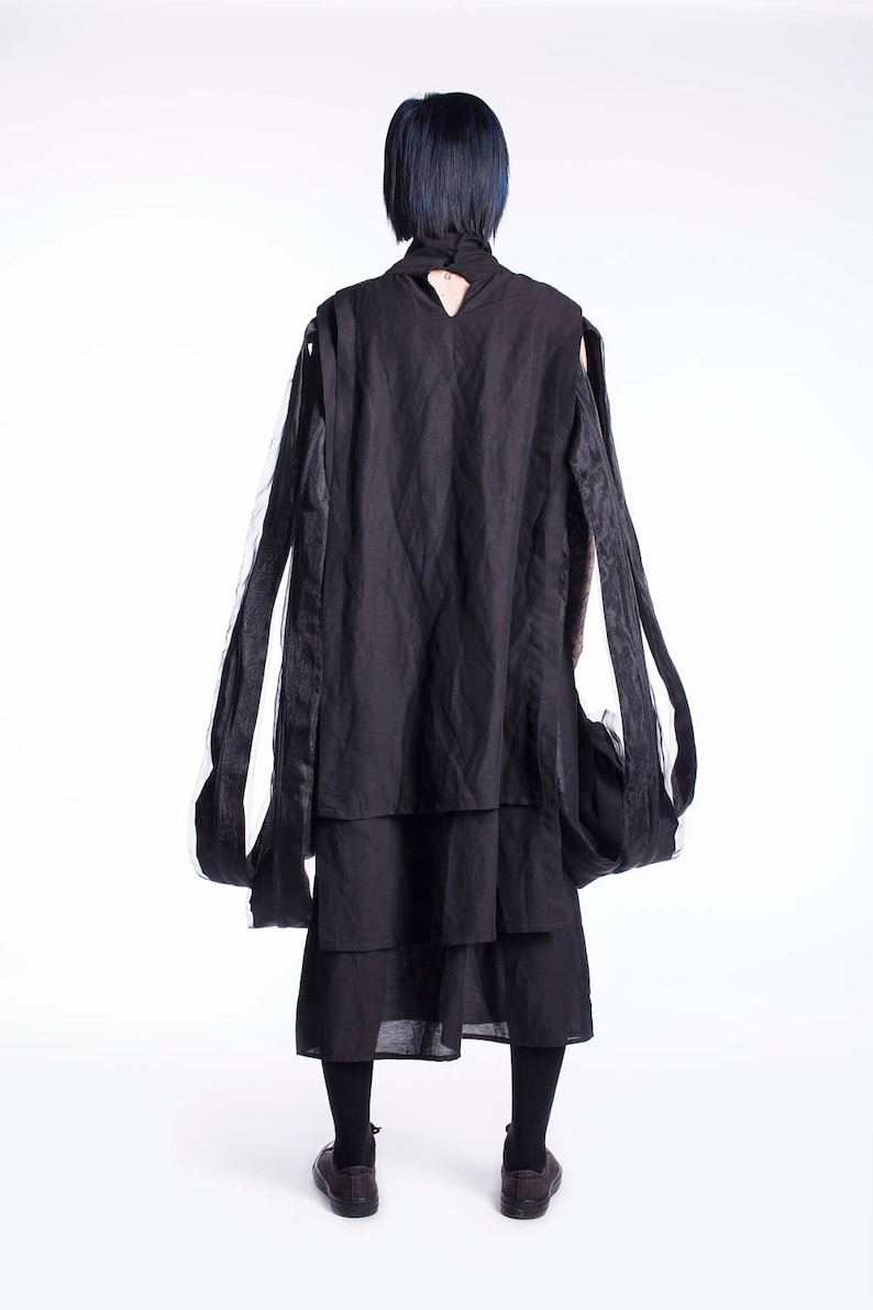 K&ouml;nnte beinhalten: Eine Person tr&auml;gt ein schwarzes, mehrschichtiges Kleid mit langen, flie&szlig;enden &Auml;rmeln. Das Kleid hat ein einzigartiges Design mit einem mehrschichtigen Rock und einem hohen Ausschnitt.