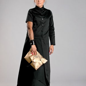 May include: A black, button-down coat dress with a high collar and long skirt. The outfit includes a gold clutch and colorful sneakers. A black leather cuff is worn on the left wrist.