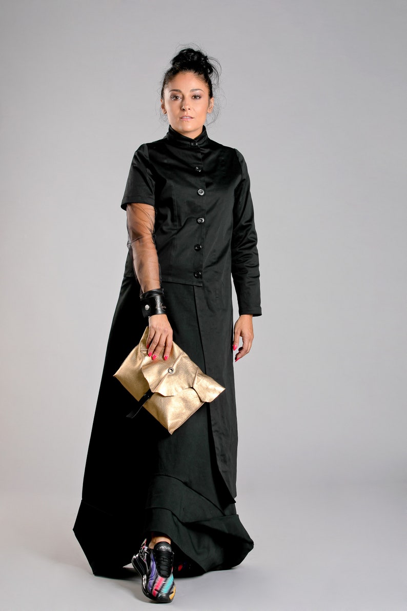 May include: A black, button-down coat dress with a high collar and long skirt. The outfit includes a gold clutch and colorful sneakers. A black leather cuff is worn on the left wrist.