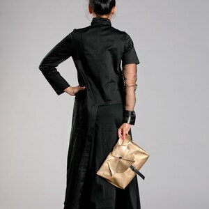May include: A woman in a black outfit, including a long coat and wide-leg pants, holds a gold clutch. The outfit has a high collar and sheer sleeves. The clutch is rectangular with a black strap.