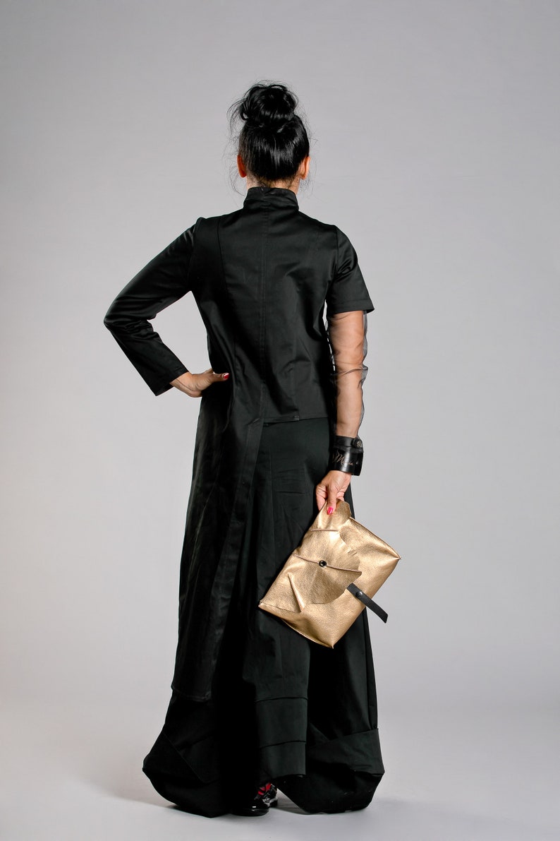May include: A woman in a black outfit, including a long coat and wide-leg pants, holds a gold clutch. The outfit has a high collar and sheer sleeves. The clutch is rectangular with a black strap.