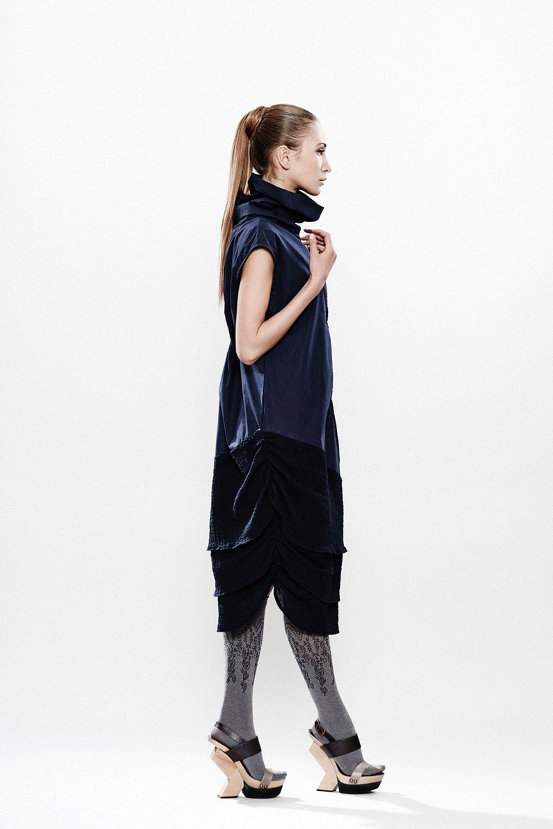 May include: A woman wearing a navy blue dress with a high neck and a ruffled skirt. She is wearing gray patterned tights and brown platform sandals.