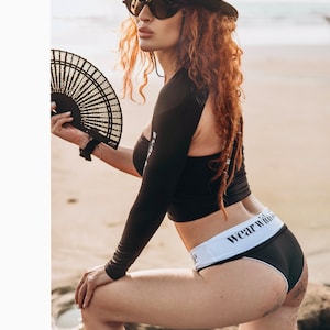 May include: A woman wearing a black one-piece swimsuit with a white waistband that says "wear wild" on it. She is wearing a black hat with feathers and sunglasses. She is holding a black hand fan.