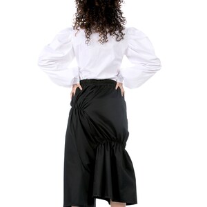 May include: A woman wearing a white blouse with puffed sleeves and a black skirt with a gathered design. She is wearing white sneakers.