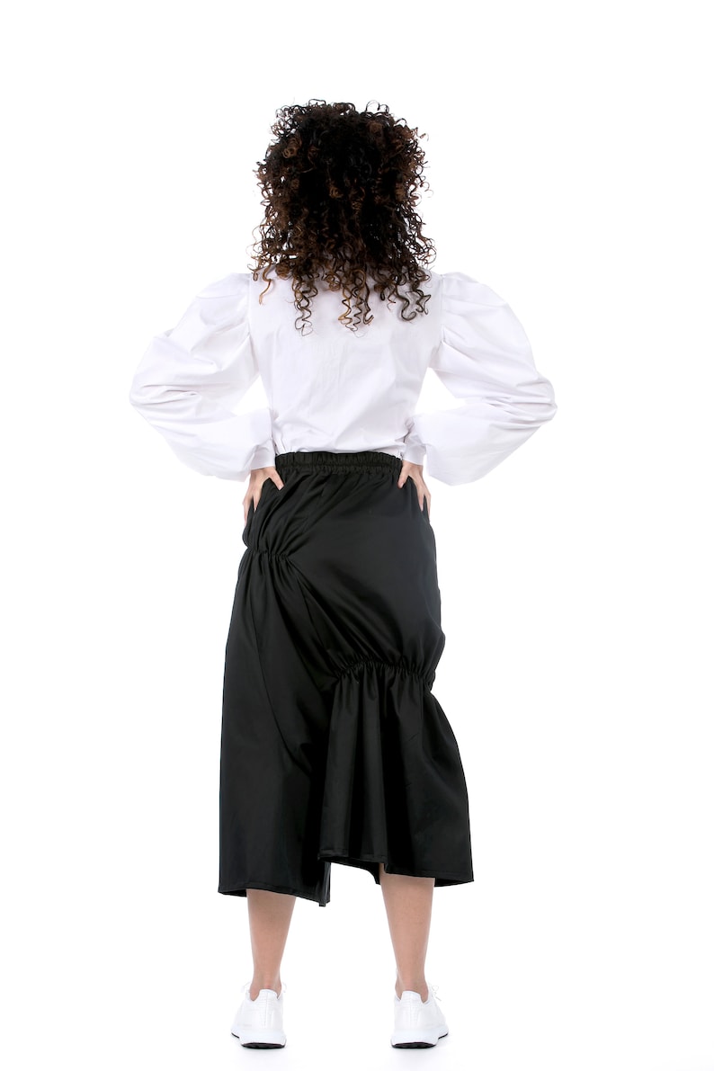 May include: A woman wearing a white blouse with puffed sleeves and a black skirt with a gathered design. She is wearing white sneakers.