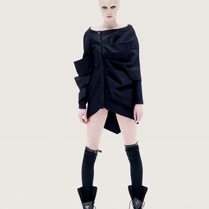 May include: A woman wearing a black, asymmetrical, long-sleeved shirt with a button closure. She is wearing black knee-high socks and black lace-up boots.
