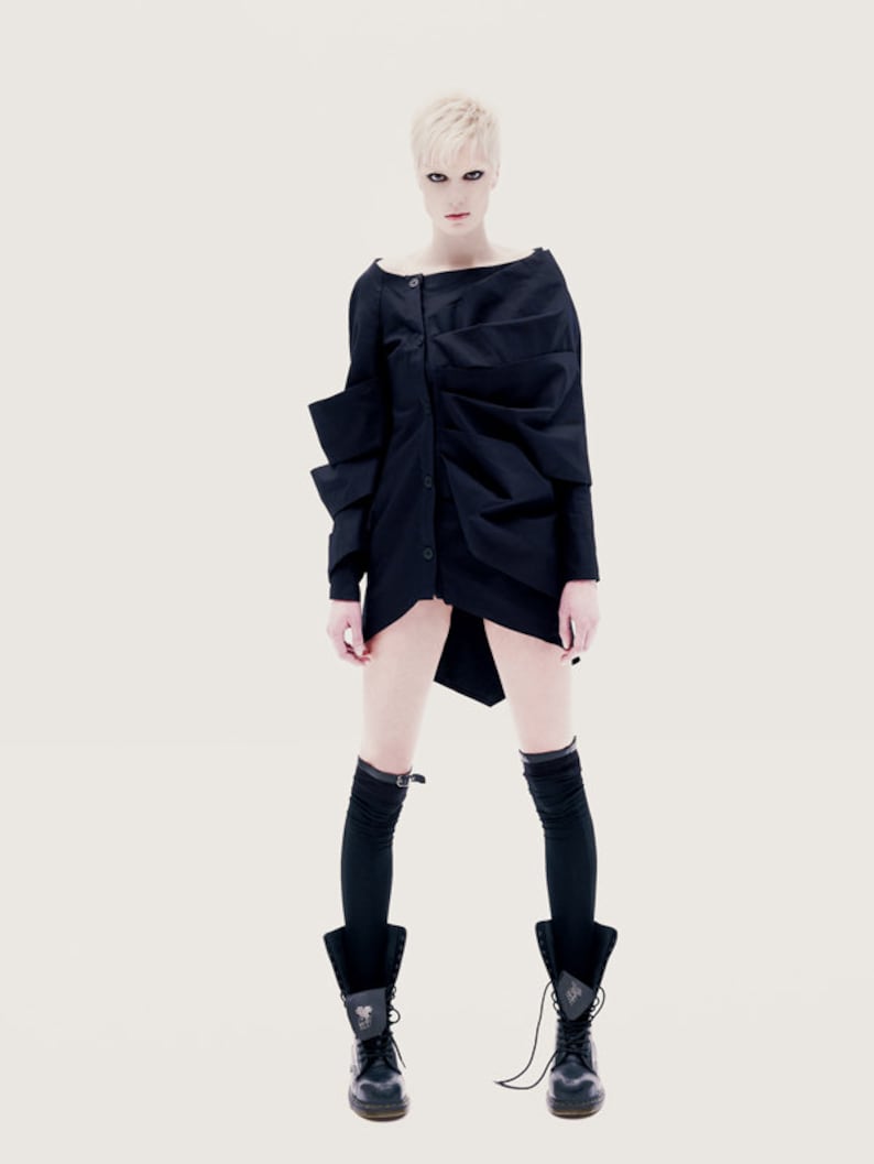 May include: A woman wearing a black, asymmetrical, long-sleeved shirt with a button closure. She is wearing black knee-high socks and black lace-up boots.
