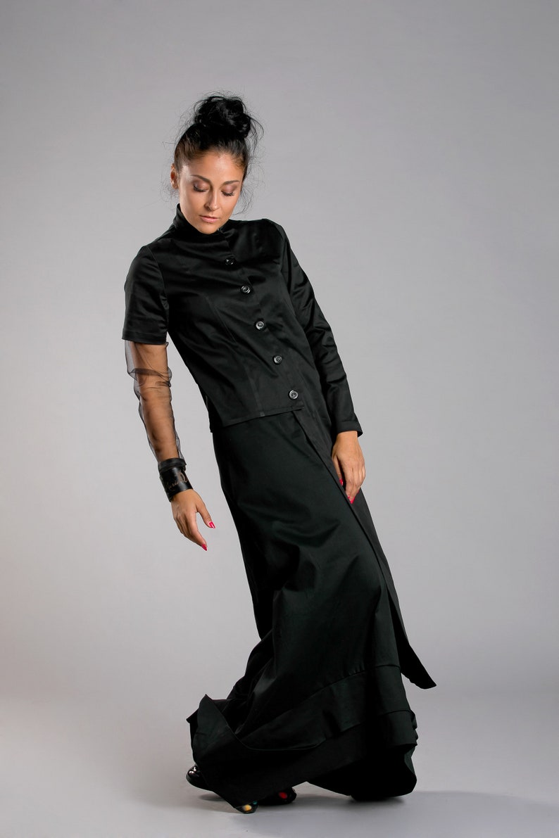May include: A black ensemble featuring a button-down top with short sleeves and a long skirt. The outfit includes a sheer sleeve detail and a wide cuff bracelet. The model is wearing black shoes with colorful accents.