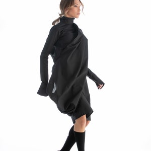 May include: A woman wearing a black, long-sleeved, asymmetrical dress with a high neck. She is wearing black socks and black lace-up shoes.