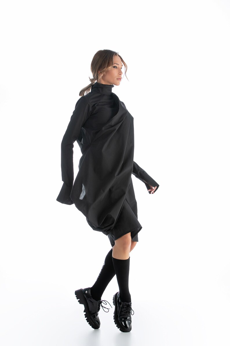 May include: A woman wearing a black, long-sleeved, asymmetrical dress with a high neck. She is wearing black socks and black lace-up shoes.